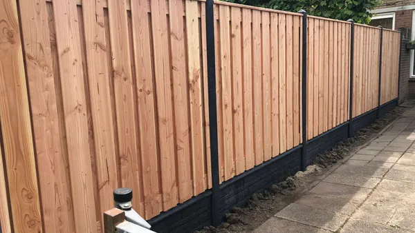 Nieuwe houten schutting met zwarte betonnen onderplaat langs tegelpad in achtertuin.