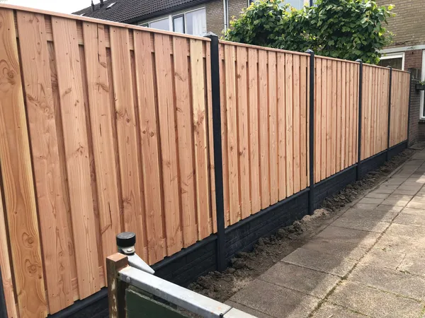 Nieuwe houten schutting met zwarte betonnen onderplaat langs tegelpad in achtertuin.