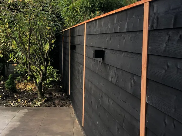 Hoge zwarte houten schutting met horizontale planken en cederhouten frame langs tegelterras.