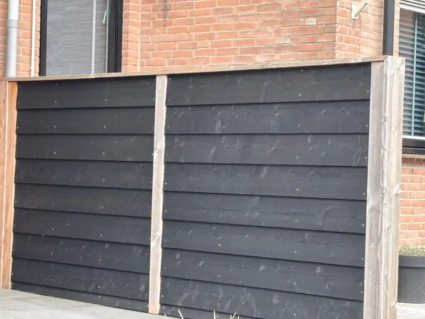 Zwarte horizontale houten schutting met cederhouten palen tegen bakstenen gevel.