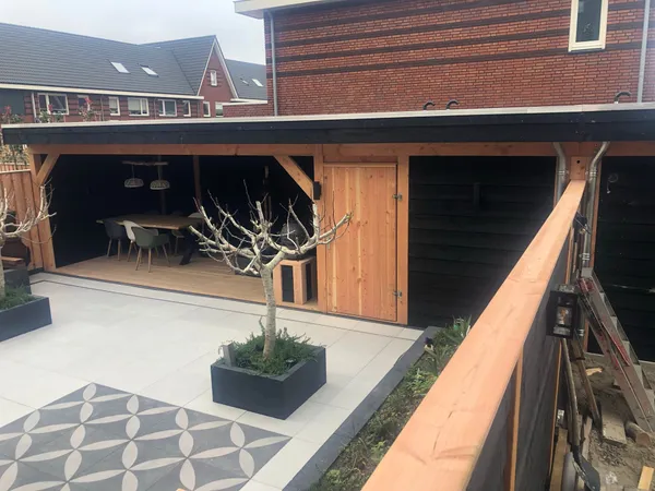 Houten overkapping met dineerruimte, aangrenzende berging en patroontegelbestrating.