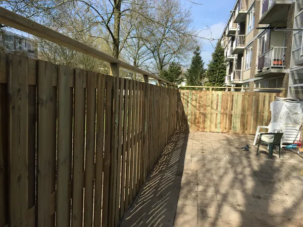Houten schutting op tegelterras bij appartementen, zijaanzicht met afdekrand.