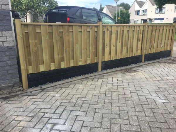 Lage houten schutting met betonnen onderplaten naast bestrate oprit.