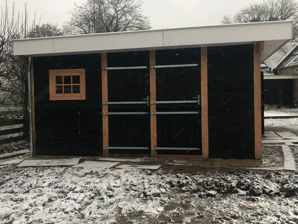 Zwarte schuur met dubbele deuren, raampje en plat dak in sneeuw.