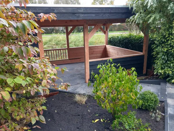 Houten overkapping in groene tuin omringd door beplanting en bestrating.